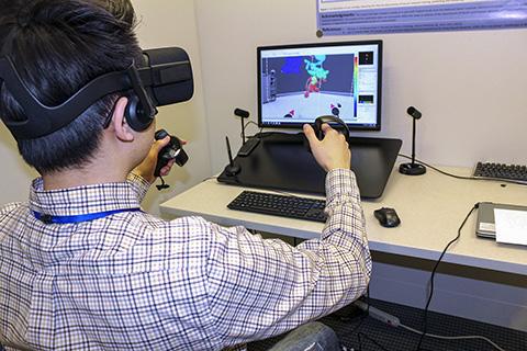 Image of a man in a plaid shirt sitting in front of a computer while wearing VR goggles and working a pair of joysticks