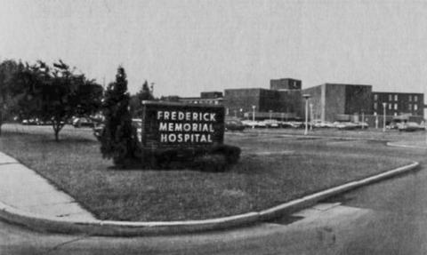 Black-and-white photo of a brick sign saying "Frederick Memorial Hospital" on a lawn, with a large brick hospital in the background