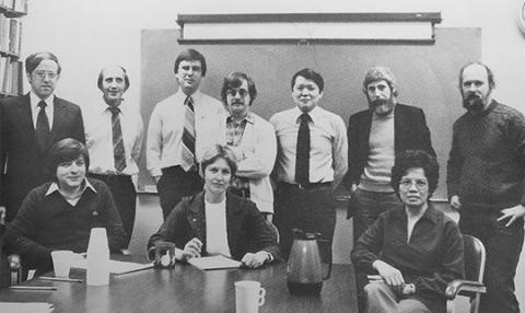 Black-and-white photo of men and women in business attire, seated and standing at a conference table