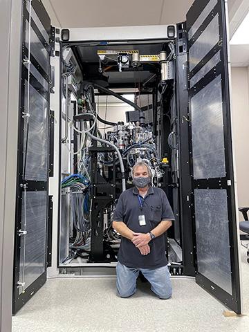 Photo of a man kneeling in front of a large, half-assembled metal and plastic cabinet containing a microscope