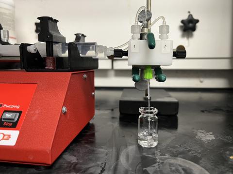 a red box machine with a start/stop button connects to vials and piping that attaches to a box with labels "1" and "2" out of which a single tube shoots liquid into a glass vial