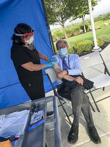 Photo of a nurse in a face mask and shield administering a vaccine to a man in business attire in a tent outdoors