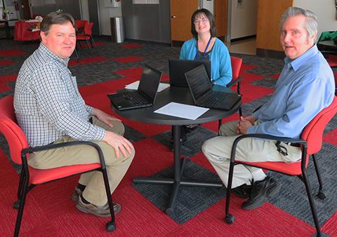 three people sit around a table with laptops