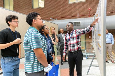 several people stand facing a person pointing at a board with a scientific poster on it