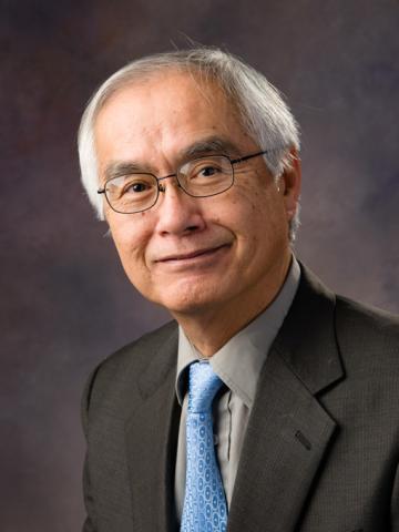 Headshot of retirement article subject, Kunio Nagashima, against a dark grey backdrop. Kunio is wearing a dark suit, gray shirt, blue necktie, and glasses.