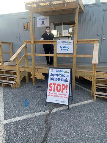 Photo of a woman wearing a surgical mask and scrubs standing on a wooden deck in front of a portable trailer