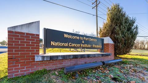 Photo of a large brick and metal sign beside a fence. The sign says, "Welcome to Fort Detrick & National Cancer Institute - Frederick"