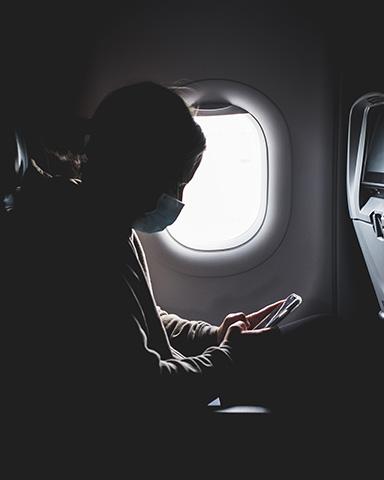 A masked person, in silhouette, looks at a cell phone beside a window on an airplane