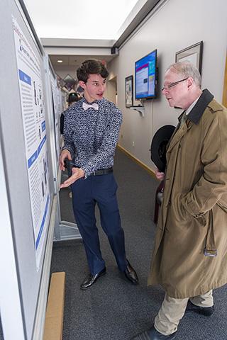 A high school student gestures to a poster hanging on a board as a senior scientist in a peacoat listens