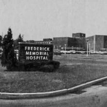 Black-and-white photo of a brick sign saying "Frederick Memorial Hospital" on a lawn, with a large brick hospital in the background