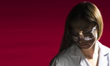 Woman with safety glasses and lab coat in front of a red background