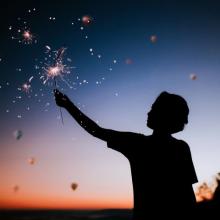 Silhouette of a young person holding a lighted sparkler against the sky at sunset