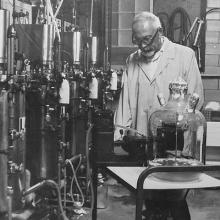 A man in a lab coat pushes a cart containing a large glass flask alongside a row of metal tanks