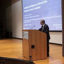 Then-Acting NCI Director Dr. Doug Lowy lectures at a podium, with a screen displaying the presentation in the background