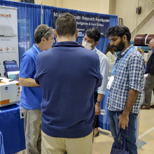 A group of men speak converse in front of a booth at an expo