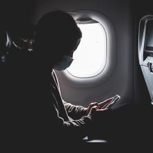 A masked person, in silhouette, looks at a cell phone beside a window on an airplane