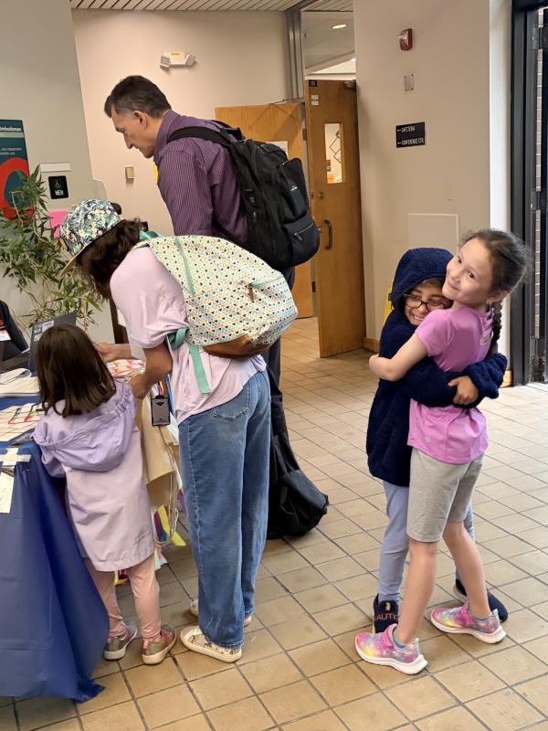 Two friends who attended a past Take Your Child to Work Day together embrace as their guardians fill out their registration at the welcome table. Image credit: Karolina Wilk