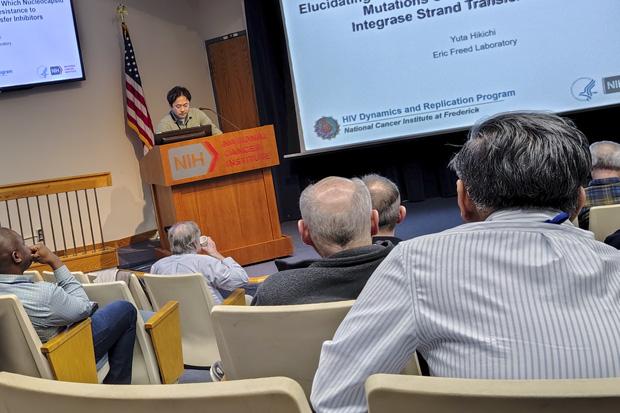 people sit facing toward a lectern with a researcher presenting along with slides on a screen